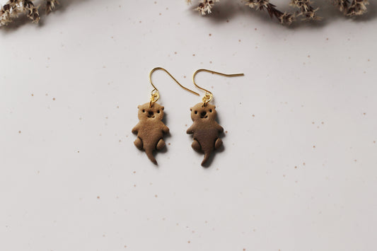 Brown otter-shaped earrings on a light gray background with dried flowers.