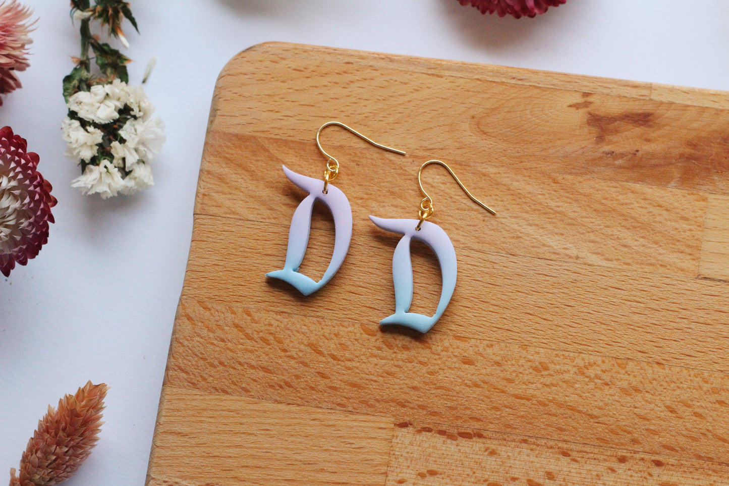 Pair of earrings with 'D' design on a wooden board with flowers in the background