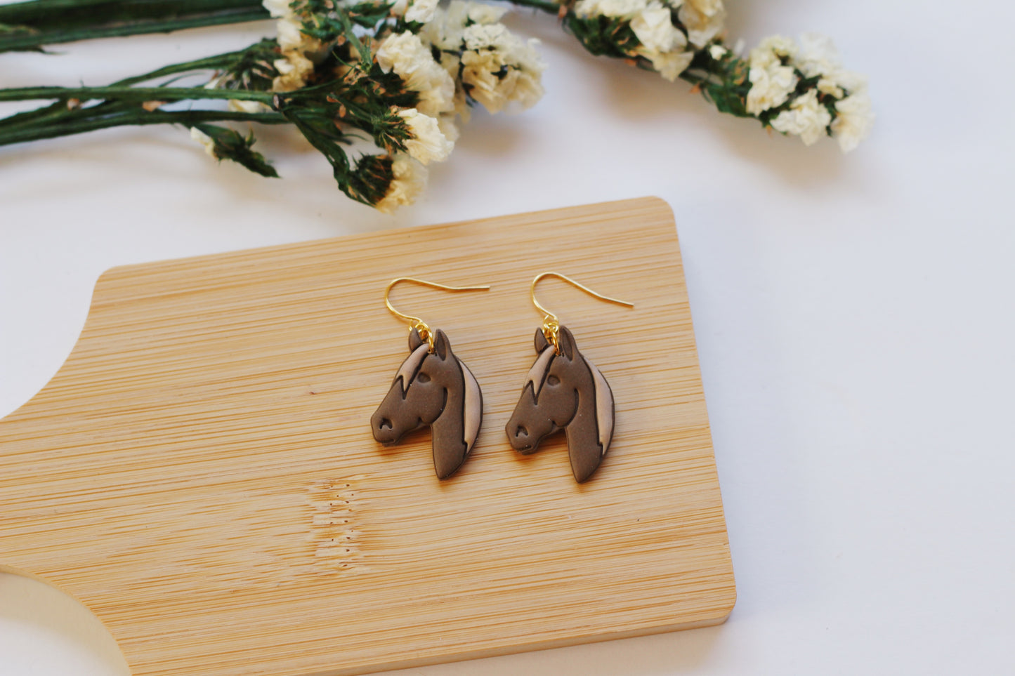 Brown horse-shaped earrings on a wooden board with flowers in the background
