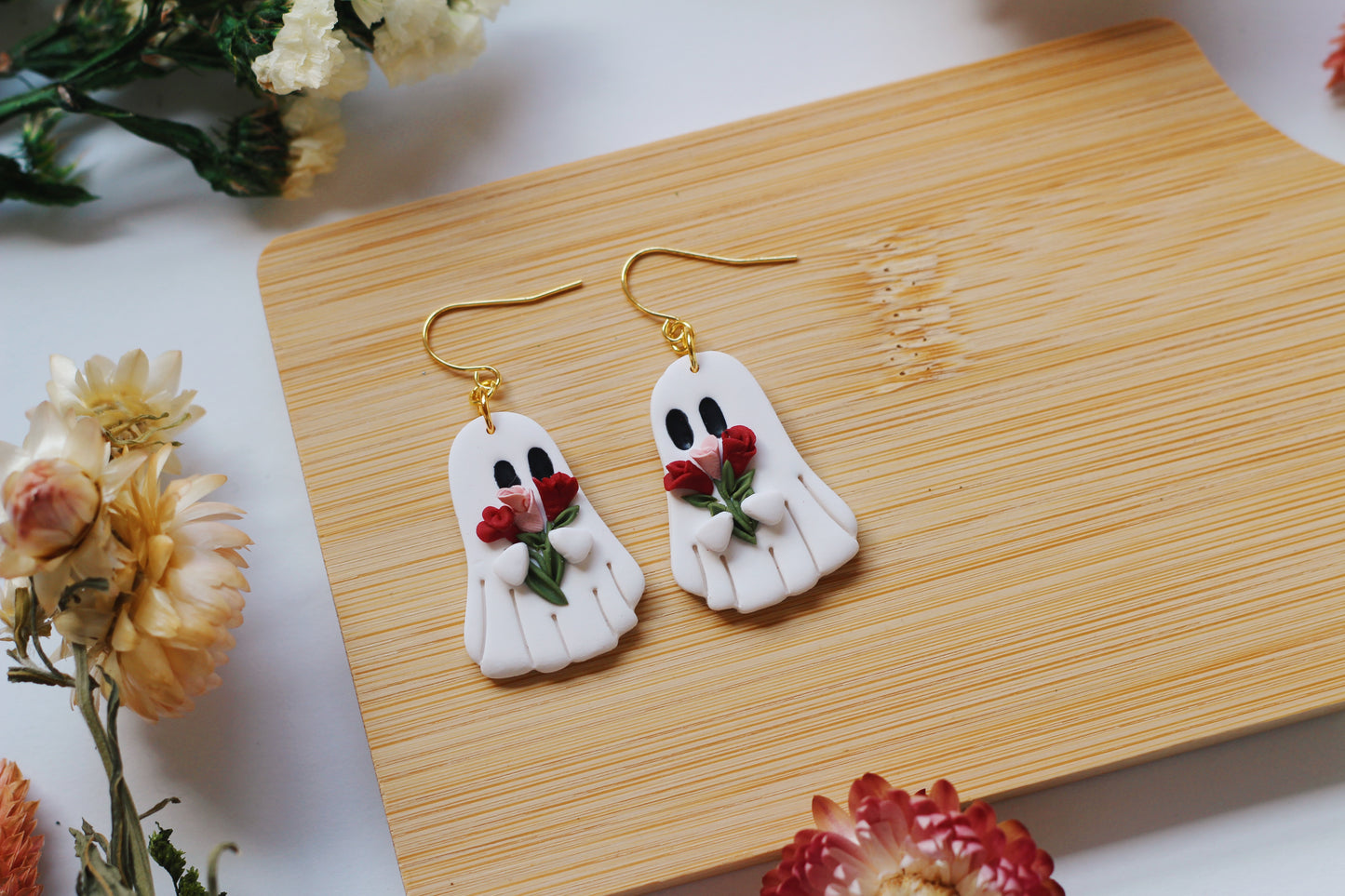 Ghost-shaped earrings holding pink and red roses on a wooden surface with flowers around