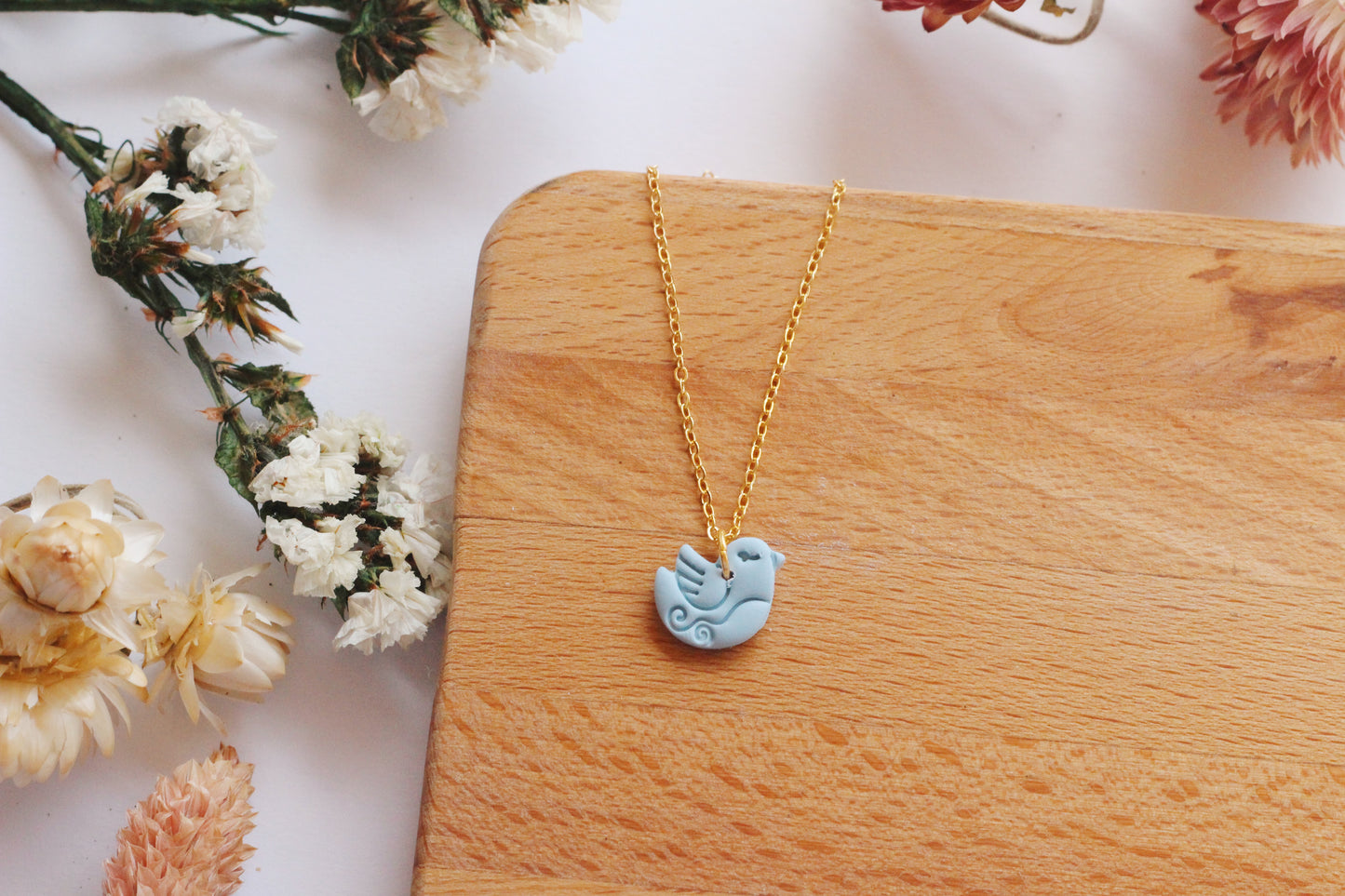 Gold necklace with a blue bird pendant on a wooden surface with flowers