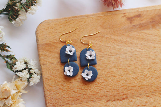 Pair of blue earrings with white flower designs on a wooden surface with flowers around.