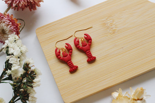 Red lobster-shaped earrings on a wooden board with flowers in the background