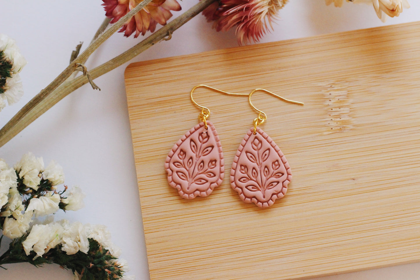 Pink teardrop earrings with gold hooks on a wooden board with flowers in the background