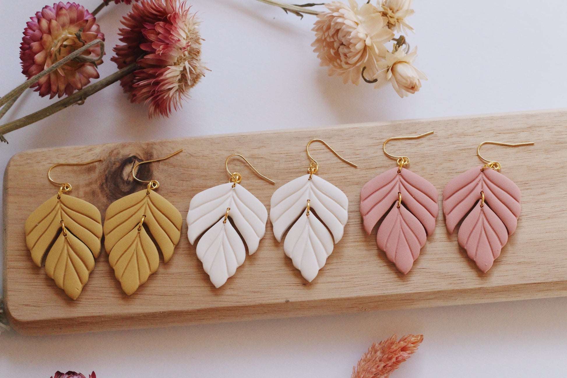 Set of leaf-shaped earrings in gold, white, and pink on a wooden board with dried flowers.