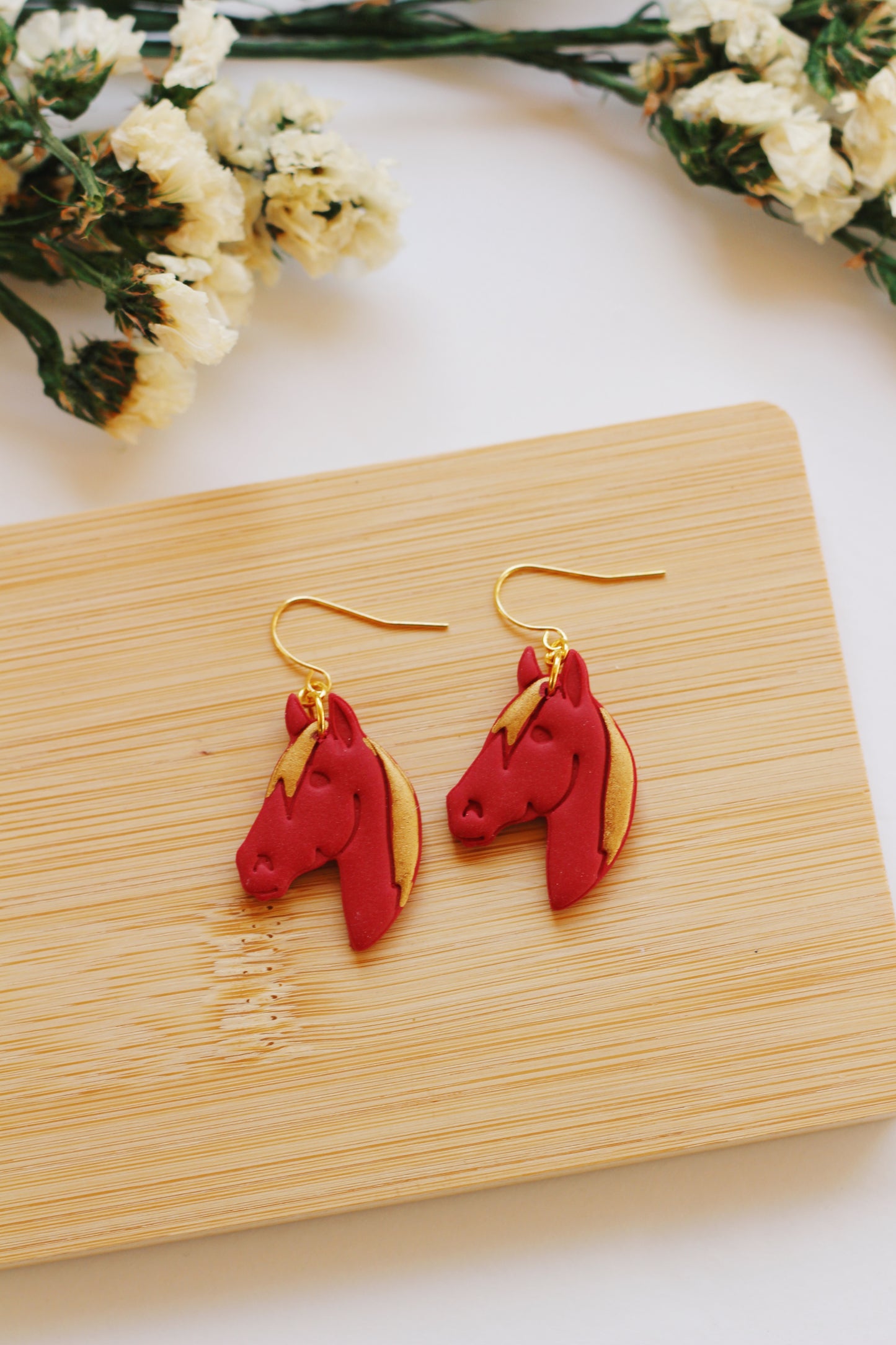 Red fire horse head earrings on a wooden board with flowers in the background