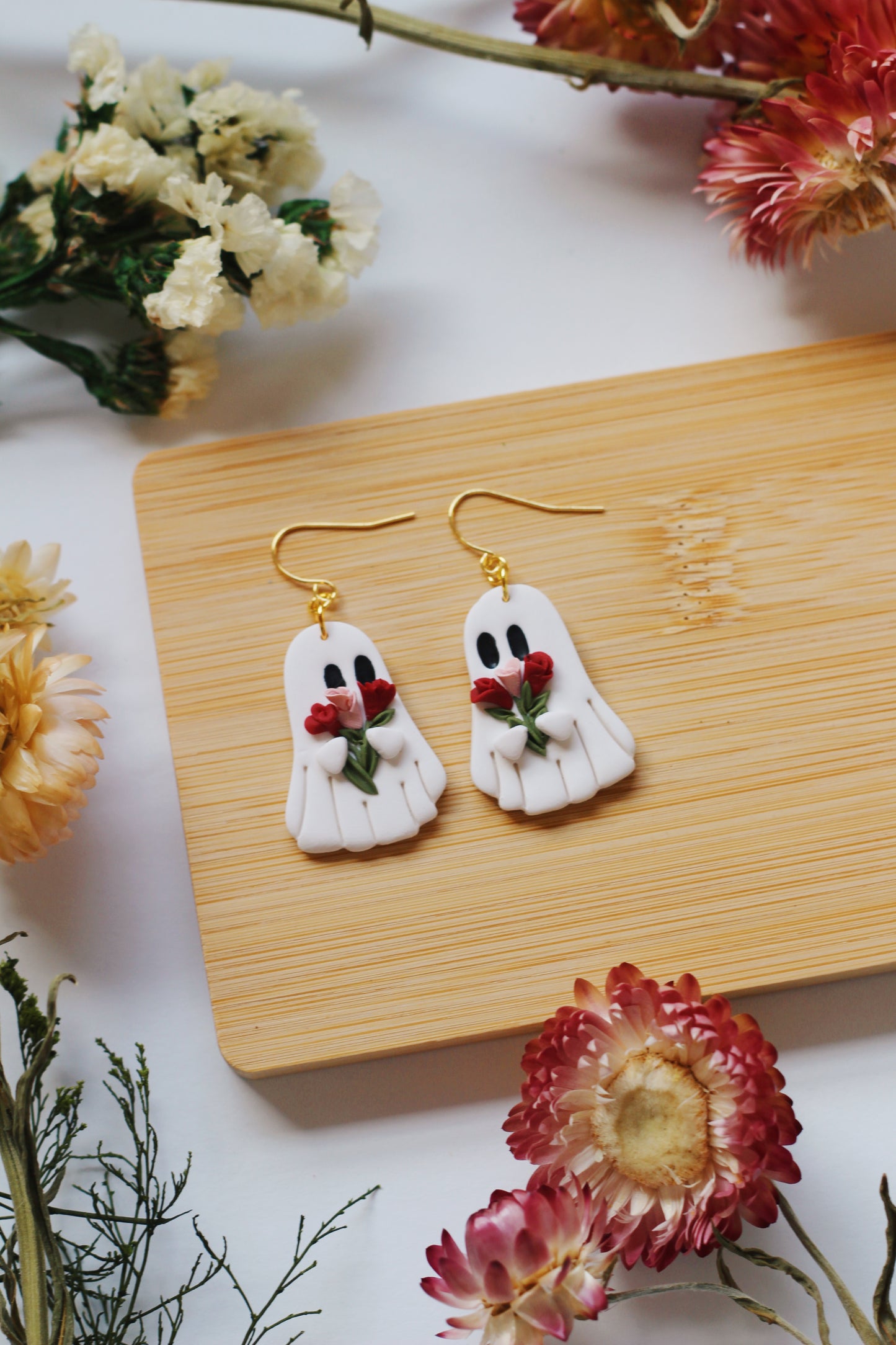 Ghost-shaped earrings with red flowers on a wooden board surrounded by flowers.