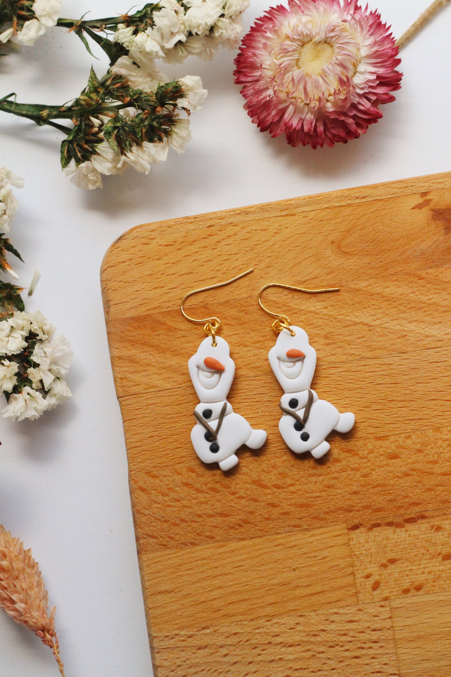 Pair of snowman-shaped earrings on a wooden surface with flowers in the background
