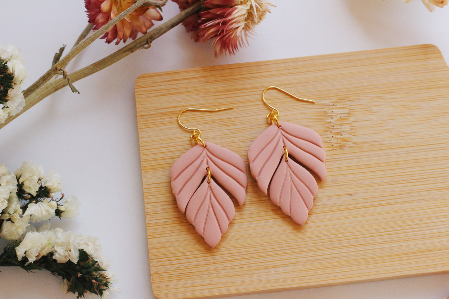 Pink leaf-shaped earrings on a wooden board with flowers in the background