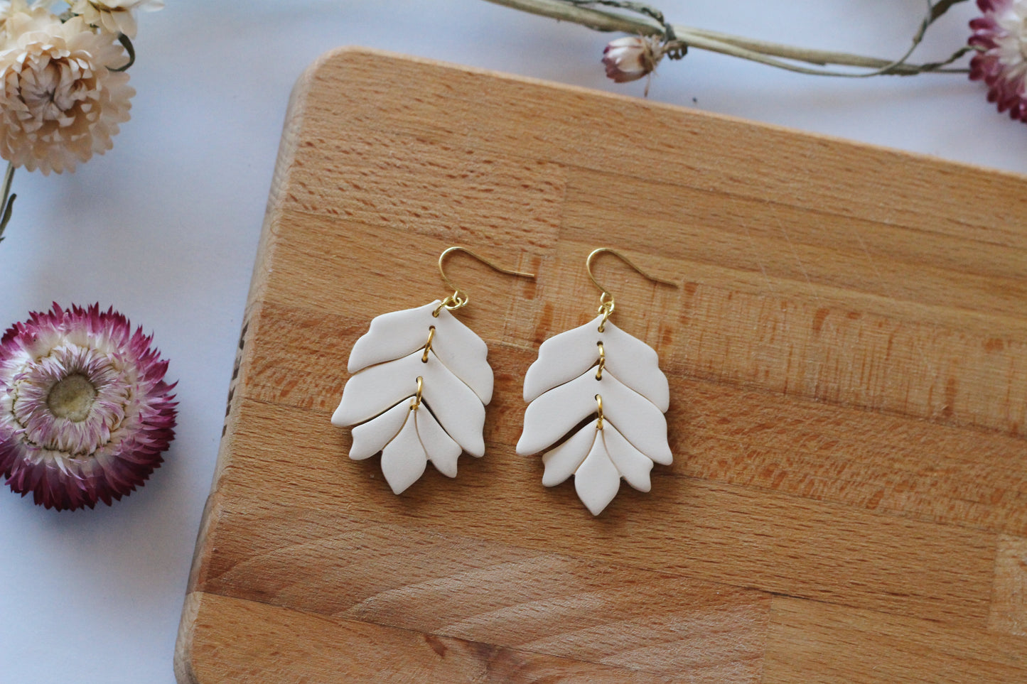 White leaf-shaped earrings on a wooden board with dried flowers.