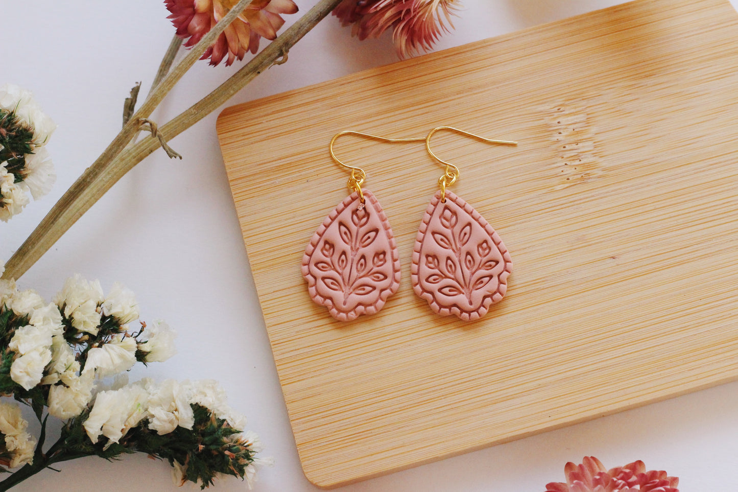 Pink teardrop earrings on a wooden board with flowers around