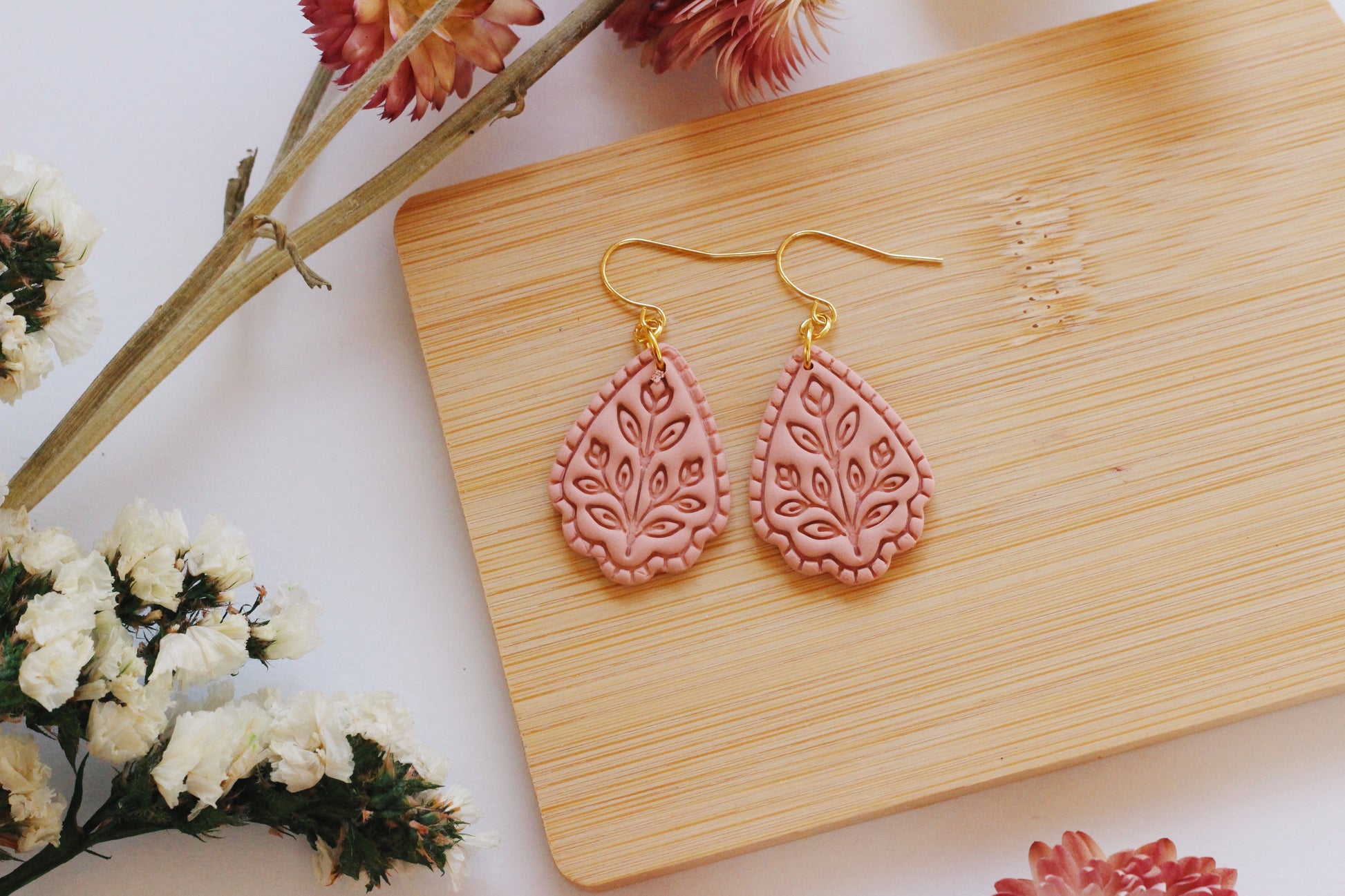 Pink teardrop earrings on a wooden board with flowers around