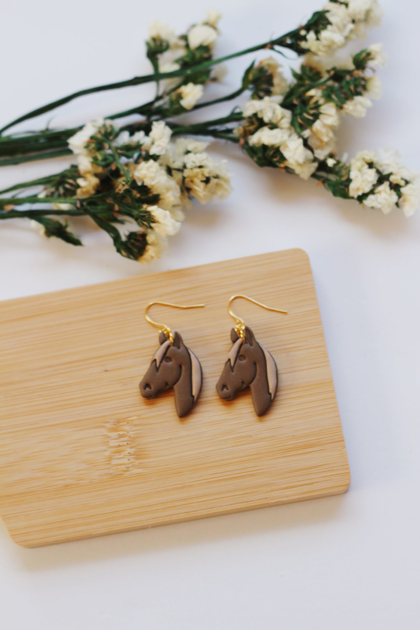 Brown horse-shaped earrings on a wooden card with white flowers in the background