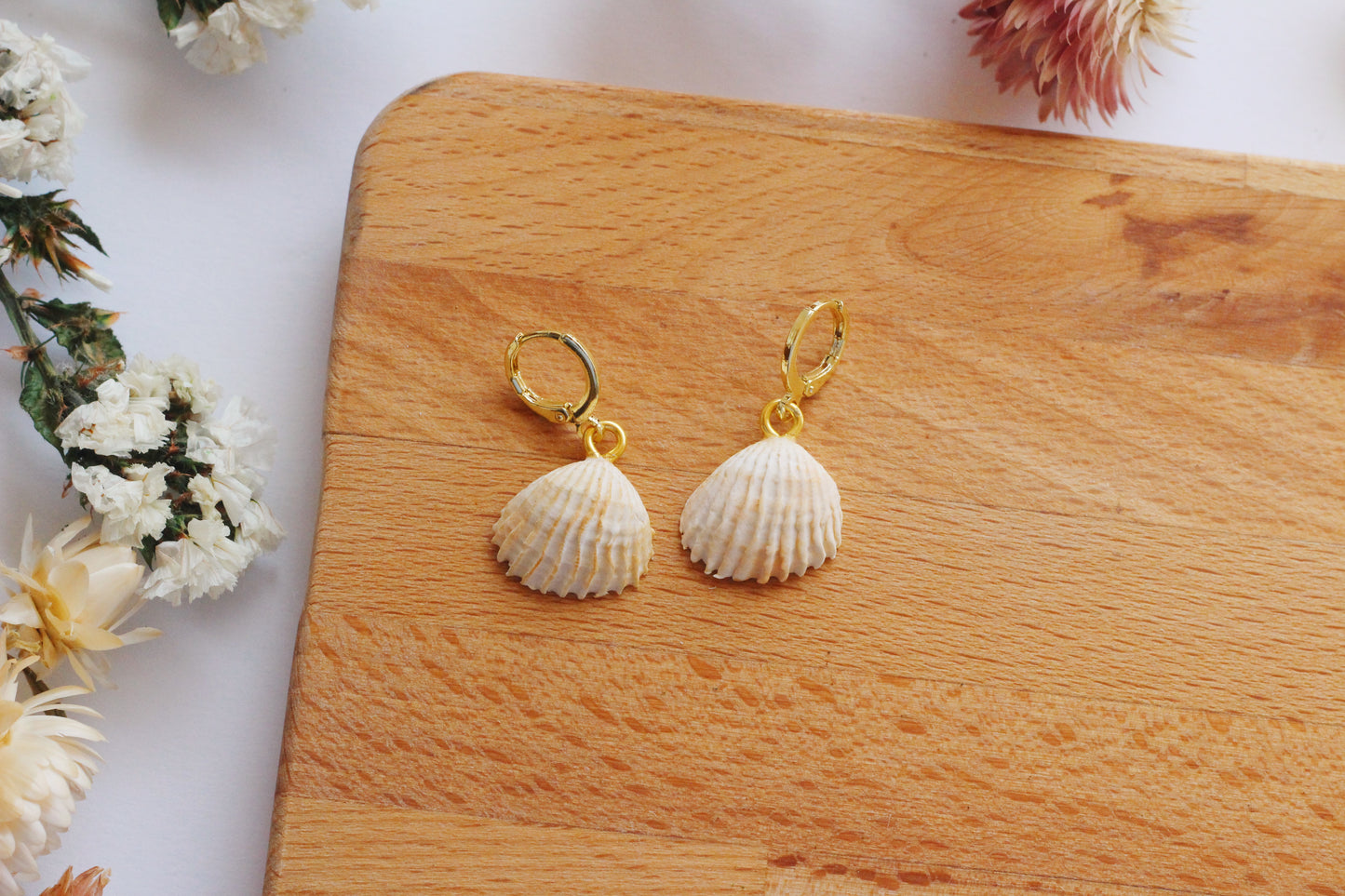 Shell-shaped earrings on a wooden board with flowers in the background