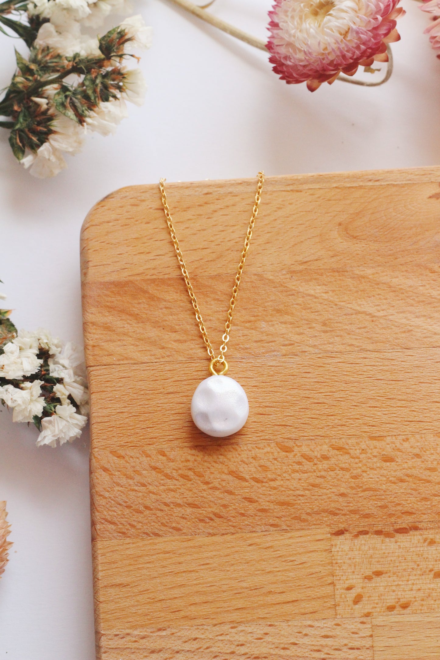 Gold necklace with a faux pearl pendant on a wooden surface with flowers in the background