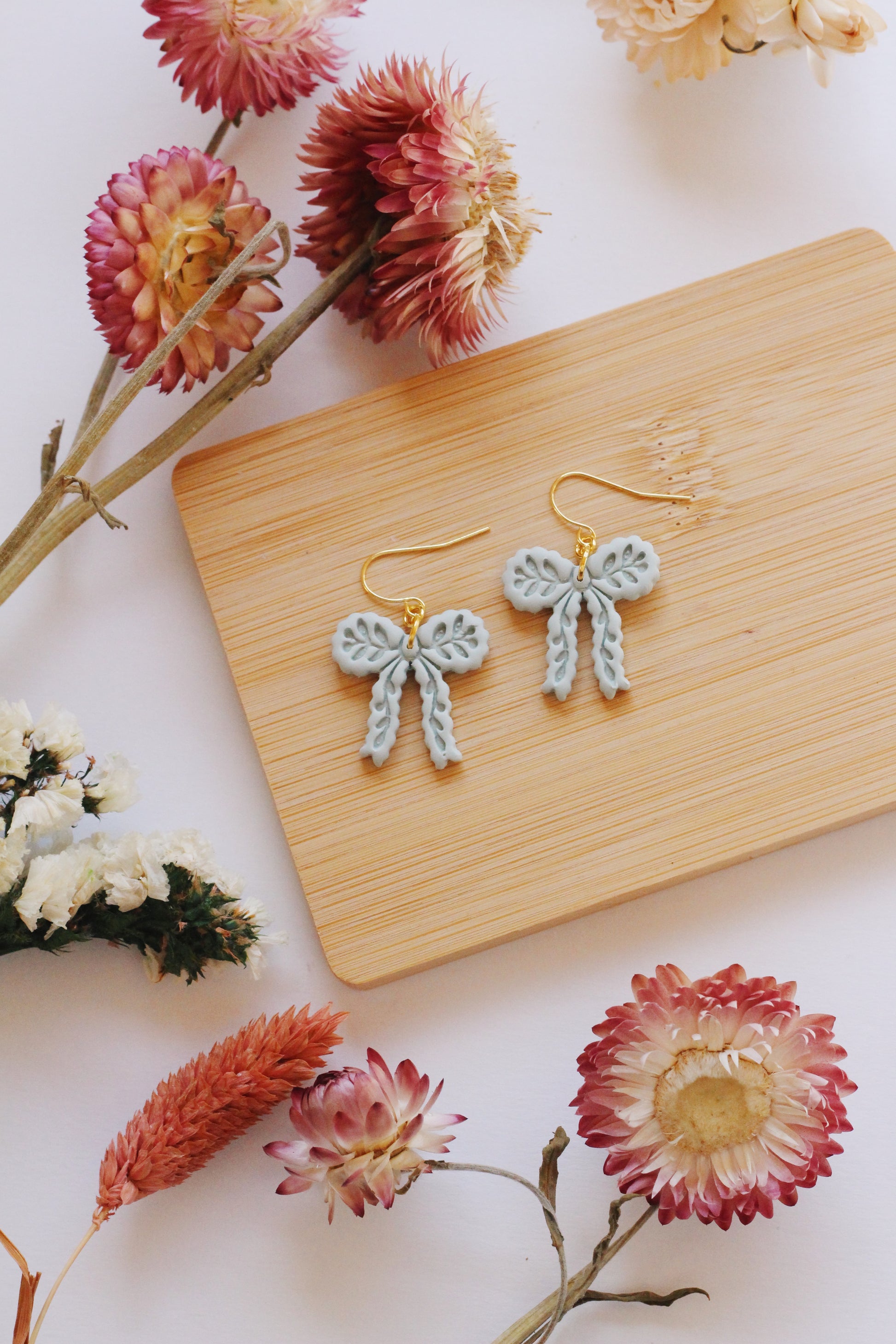 Bow-shaped earrings on a wooden board with flowers around