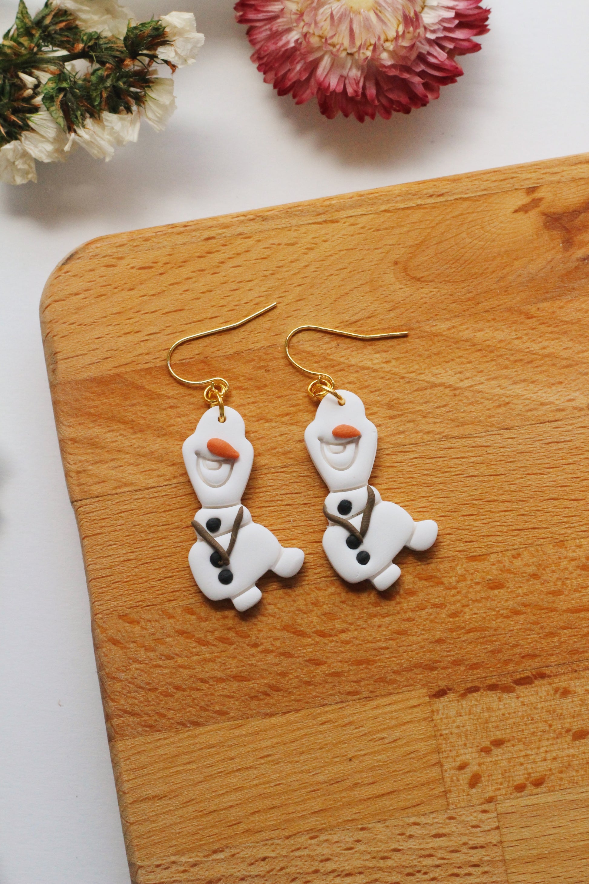 Snowman-shaped earrings on a wooden board with flowers in the background