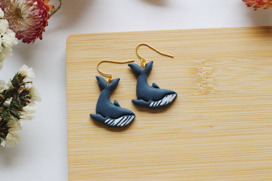 Blue whale-shaped earrings on a wooden board with flowers in the background