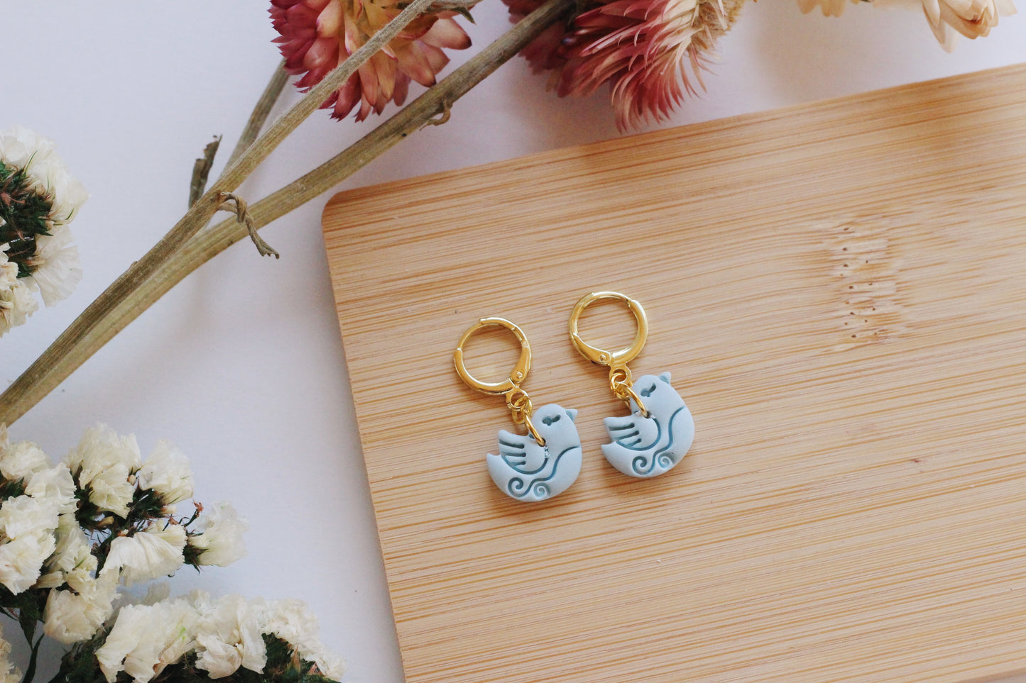 Pair of earrings with bird design on a wooden board with flowers in the background
