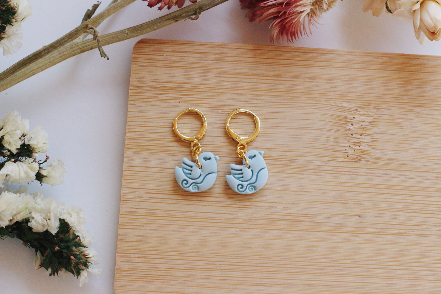 Pair of earrings with bird design on a wooden surface with flowers in the background