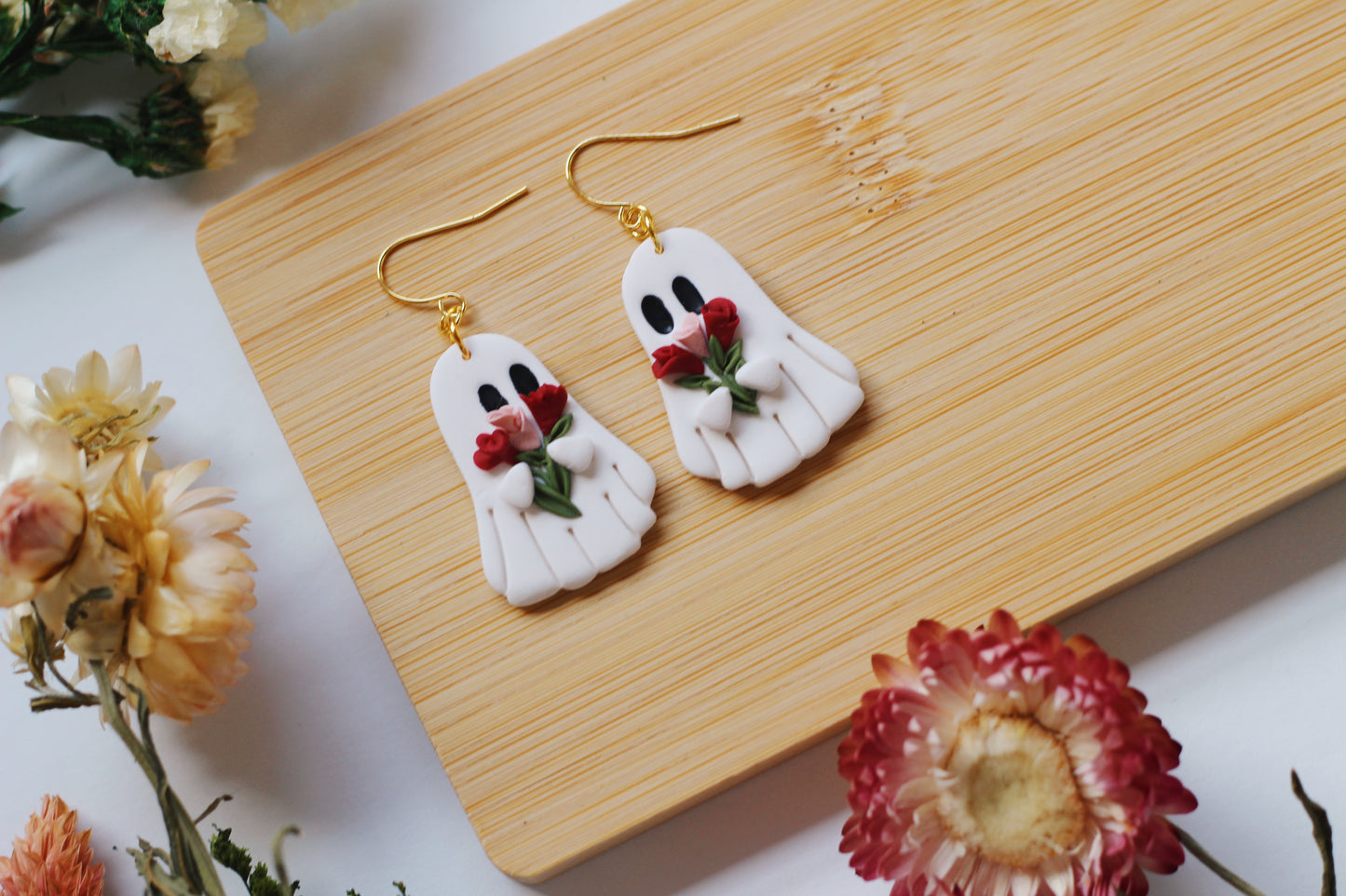 Ghost-shaped earrings with red and pink roses on a wooden board surrounded by flowers