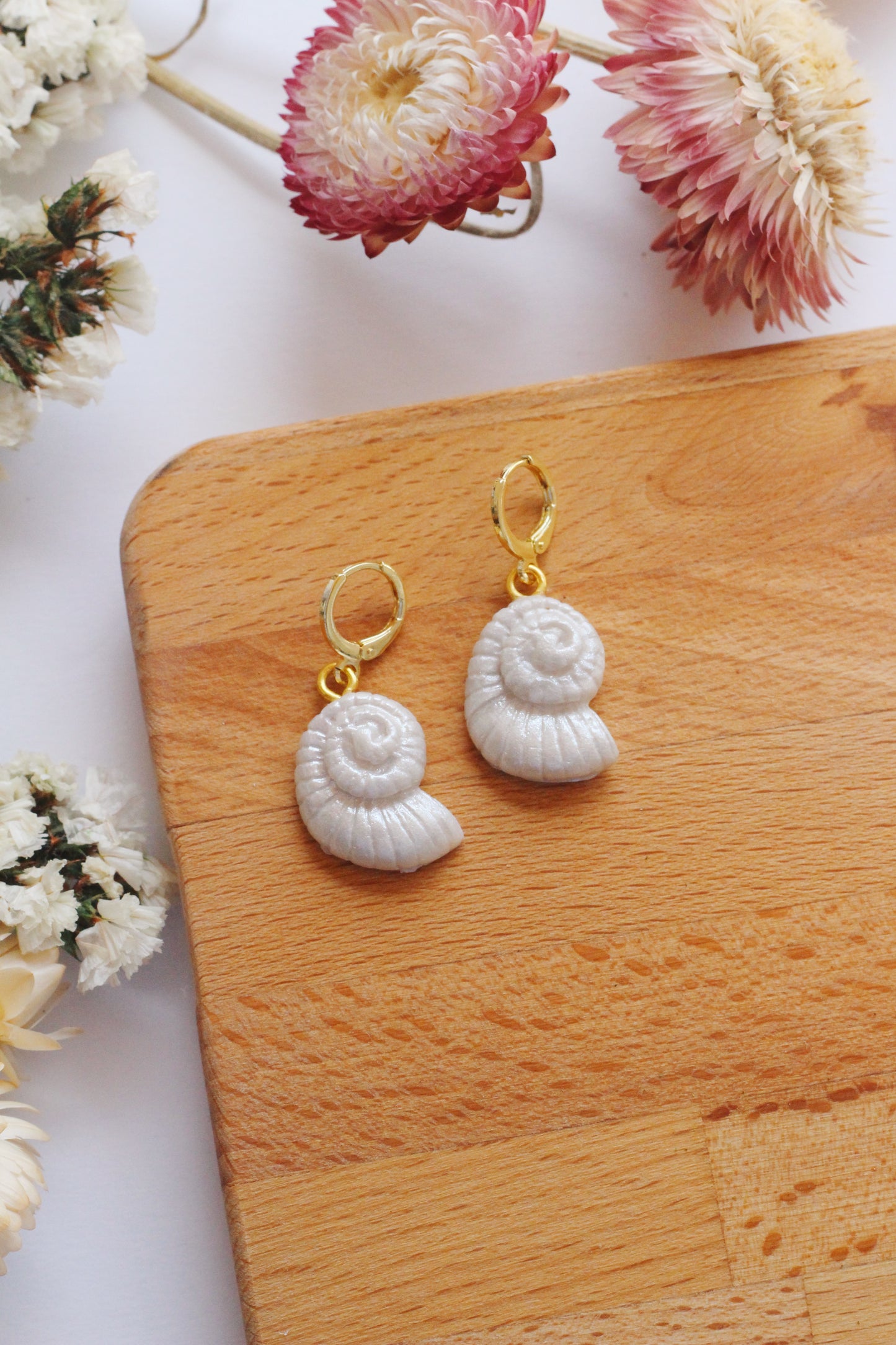 Shell-shaped earrings on a wooden board with flowers in the background