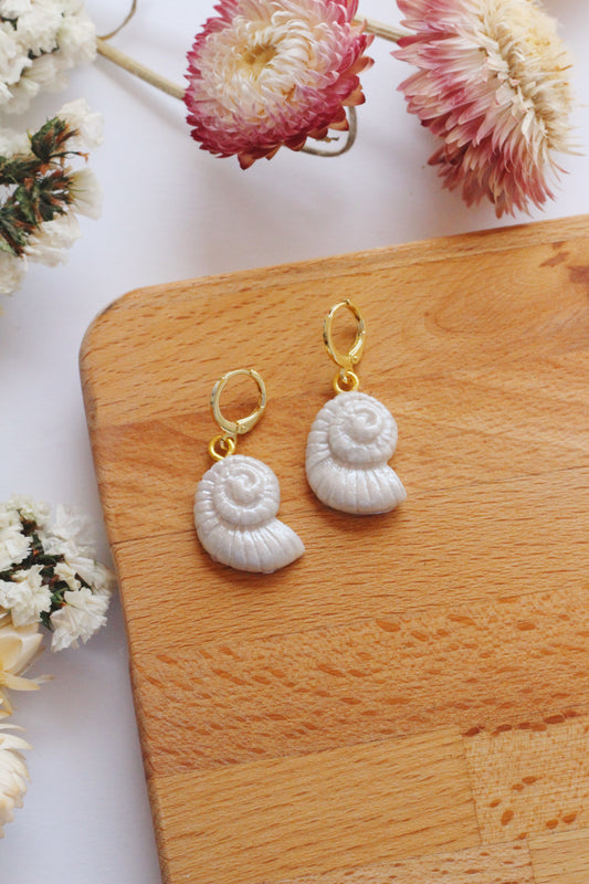 Shell-shaped earrings on a wooden board with flowers in the background