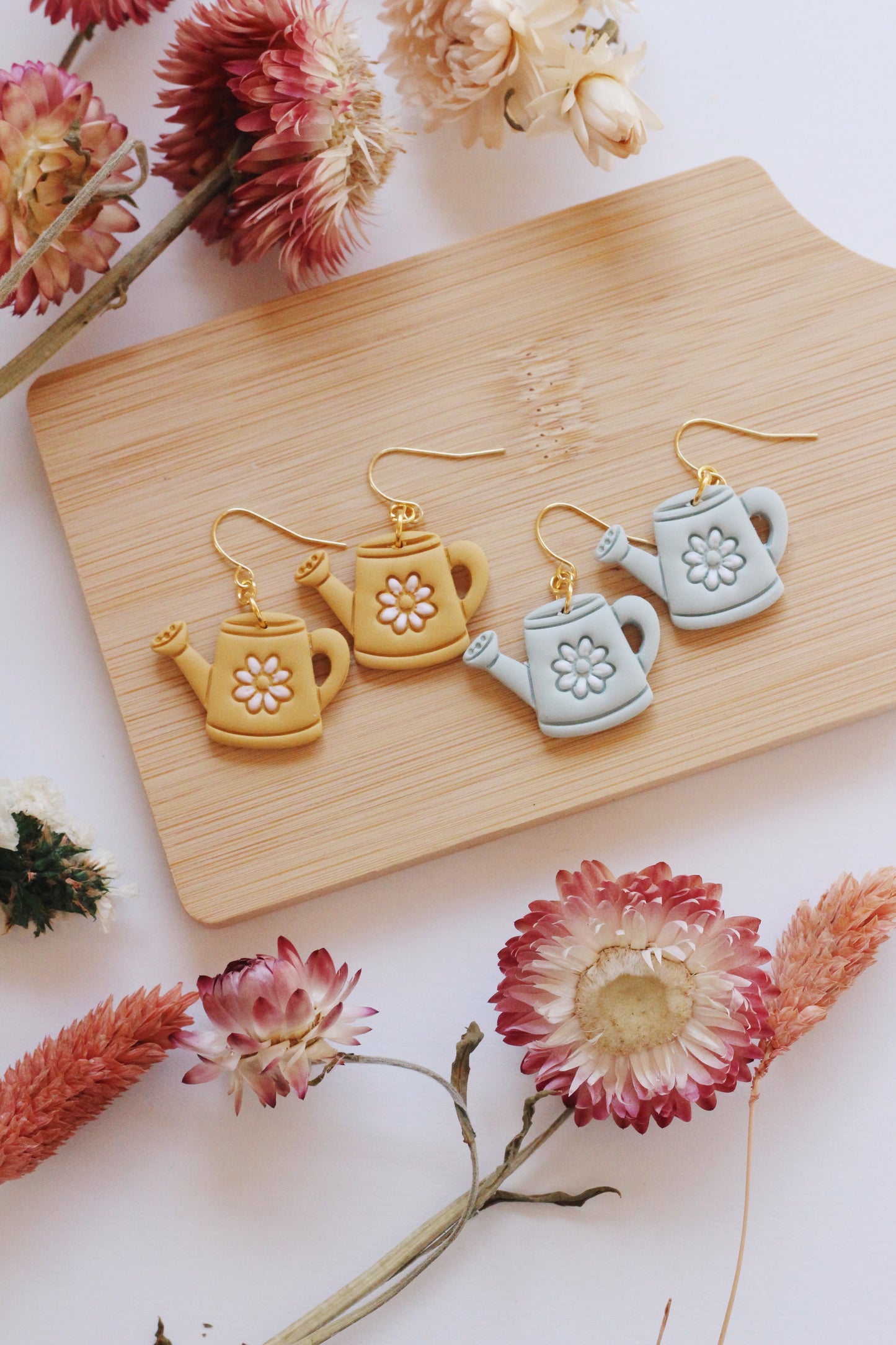 Set of ceramic watering can earrings on a wooden board with flowers around.