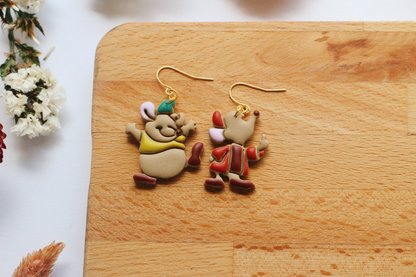 Two cartoon-style earrings on a wooden board with flowers in the background