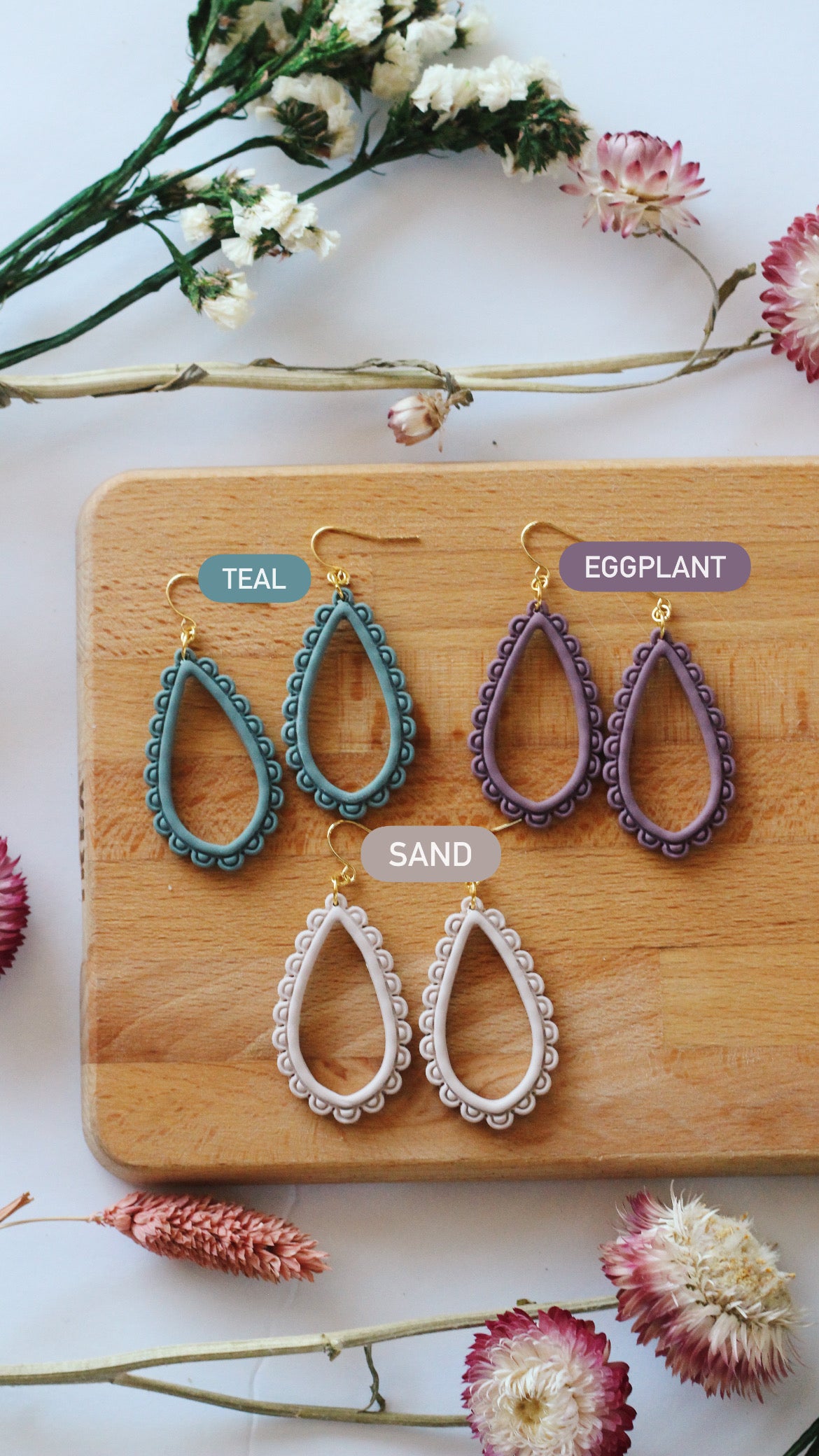 Three pairs of earrings in teal, eggplant, and sand on a wooden board with flowers around.