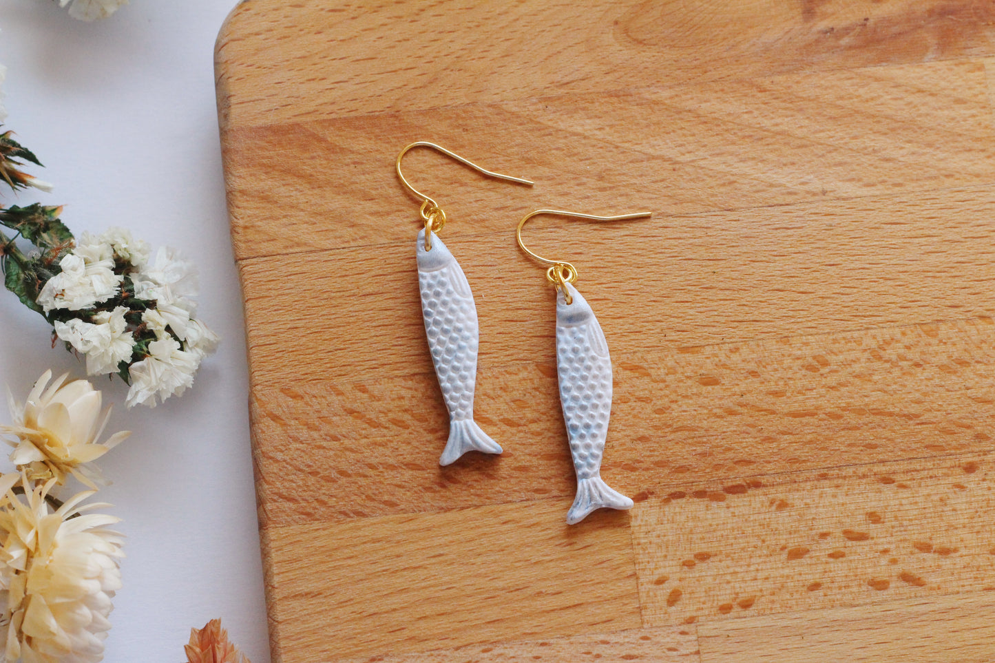 Sardine-shaped earrings on a wooden surface with flowers in the background