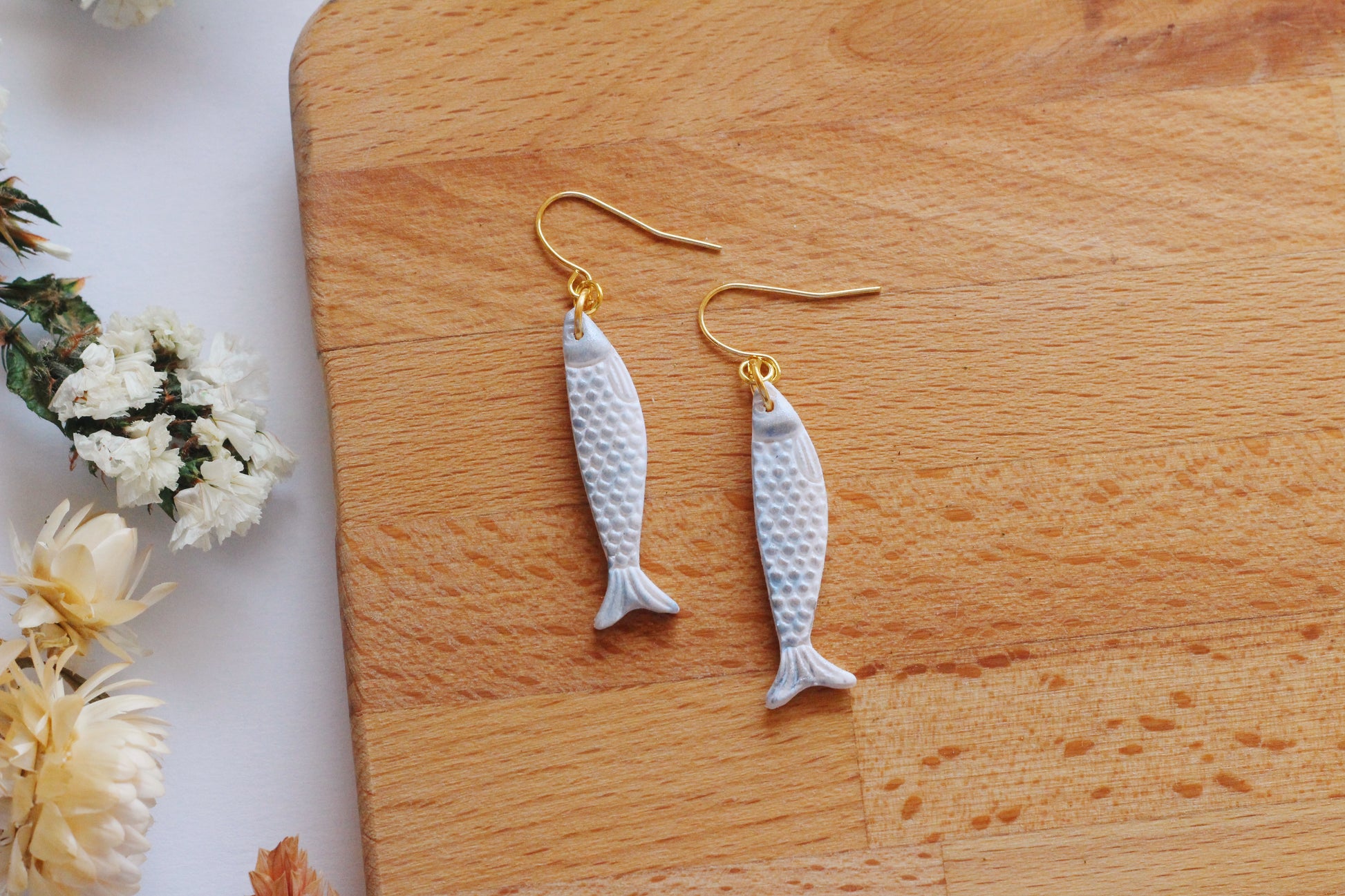 Sardine-shaped earrings on a wooden surface with flowers in the background