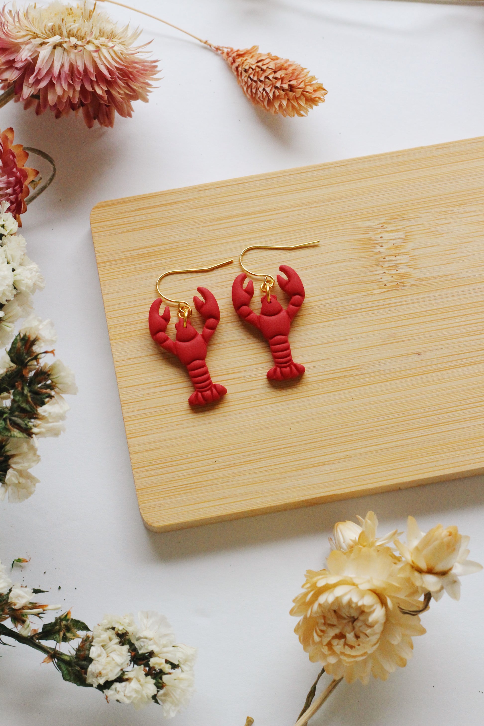 Red lobster-shaped earrings on a wooden board with flowers around