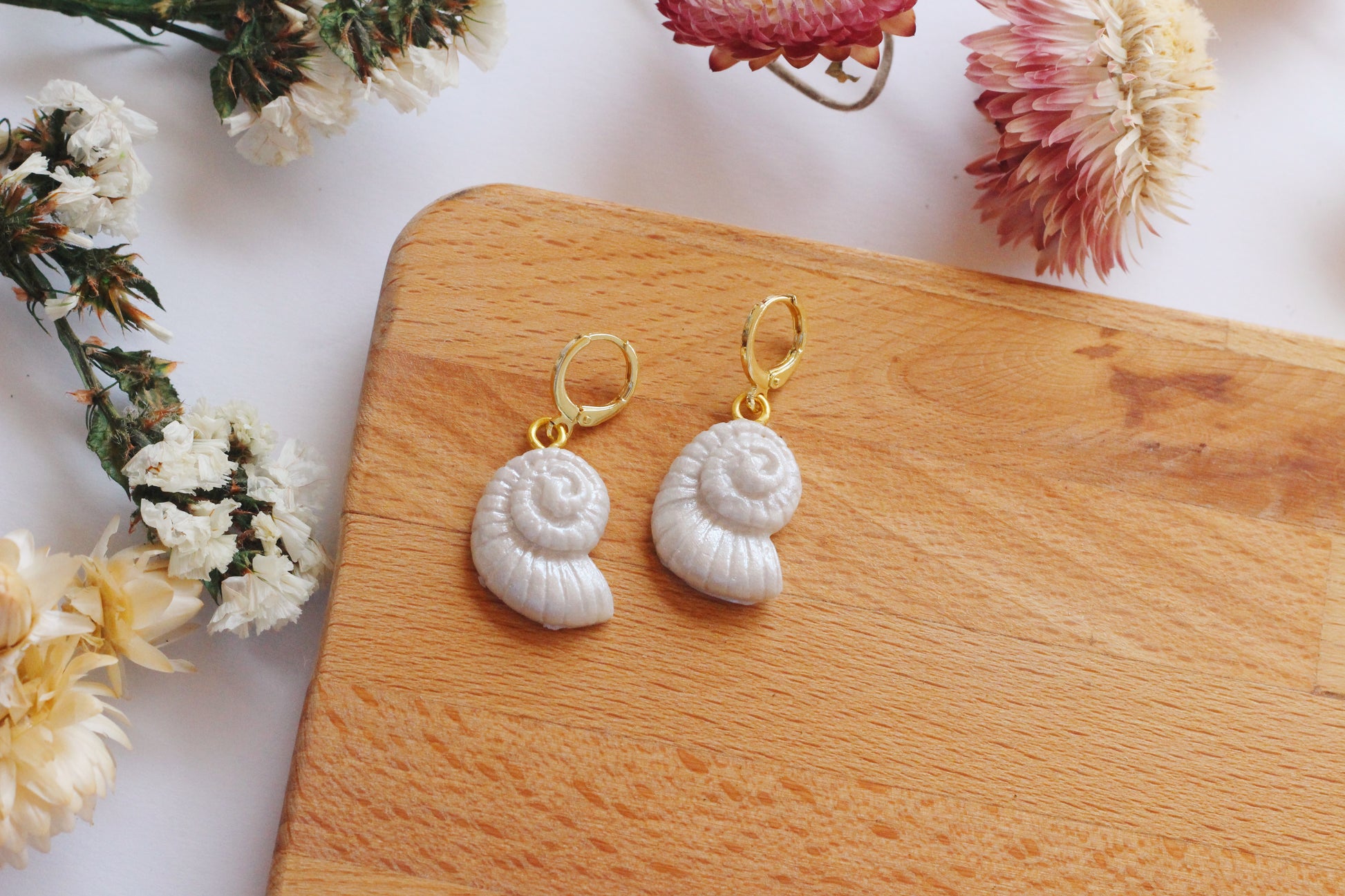 Shell-shaped earrings on a wooden board with flowers in the background