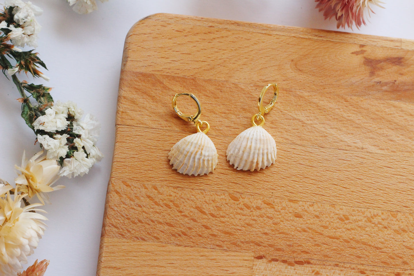 Shell-shaped earrings on a wooden board with flowers in the background