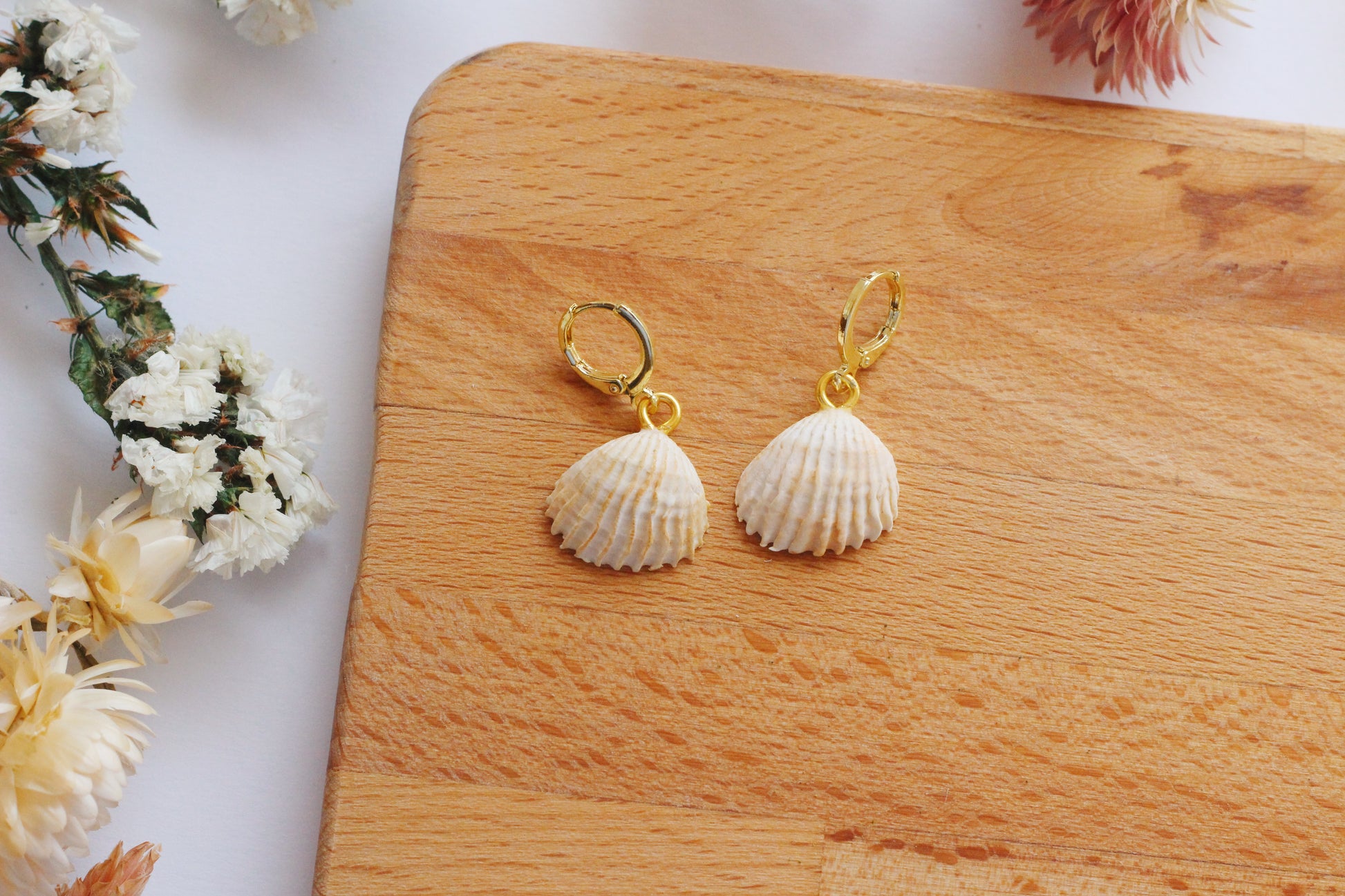 Shell-shaped earrings on a wooden board with flowers in the background