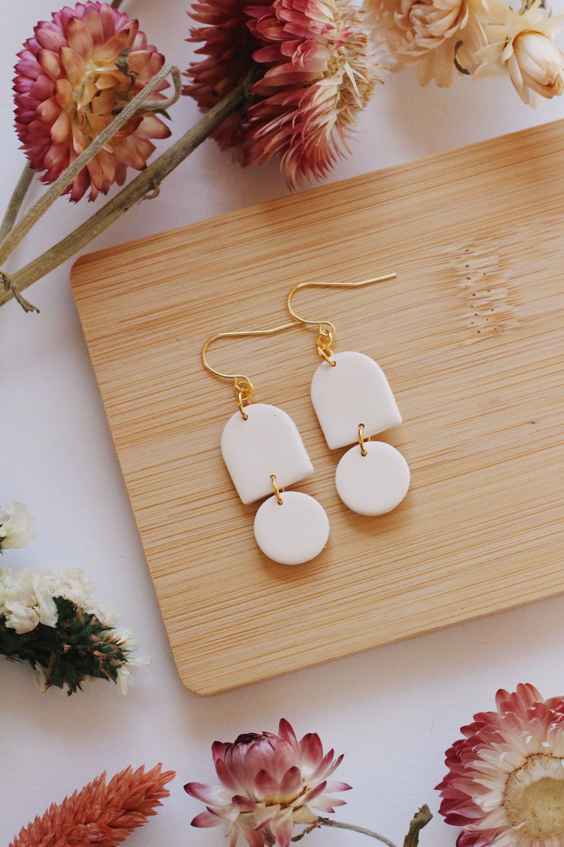 White earrings with gold hooks on a wooden board surrounded by flowers