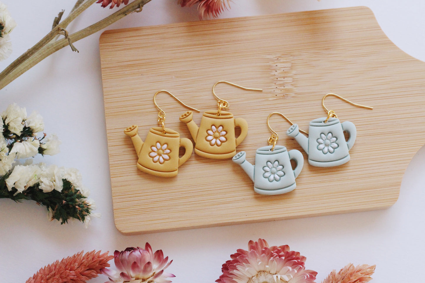 Set of watering can-shaped earrings on a wooden board with flowers around.