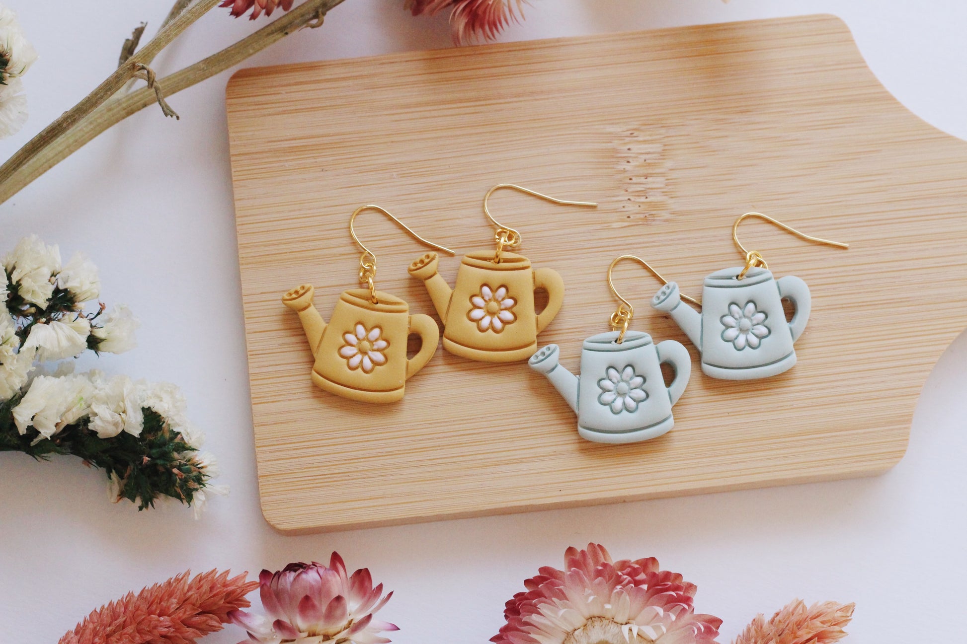 Set of watering can-shaped earrings on a wooden board with flowers around.