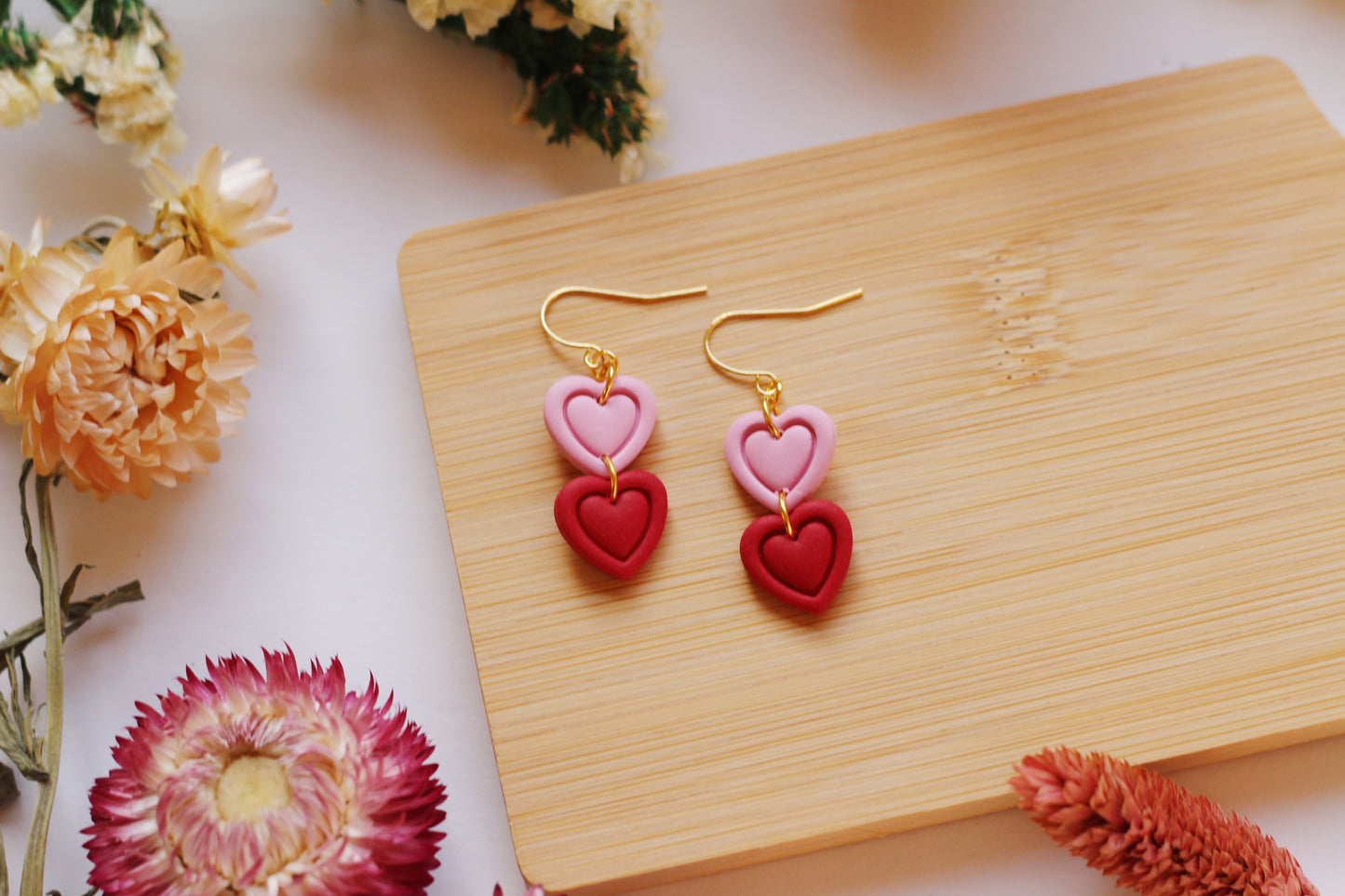 Pink and red heart-shaped earrings on a wooden board with flowers around
