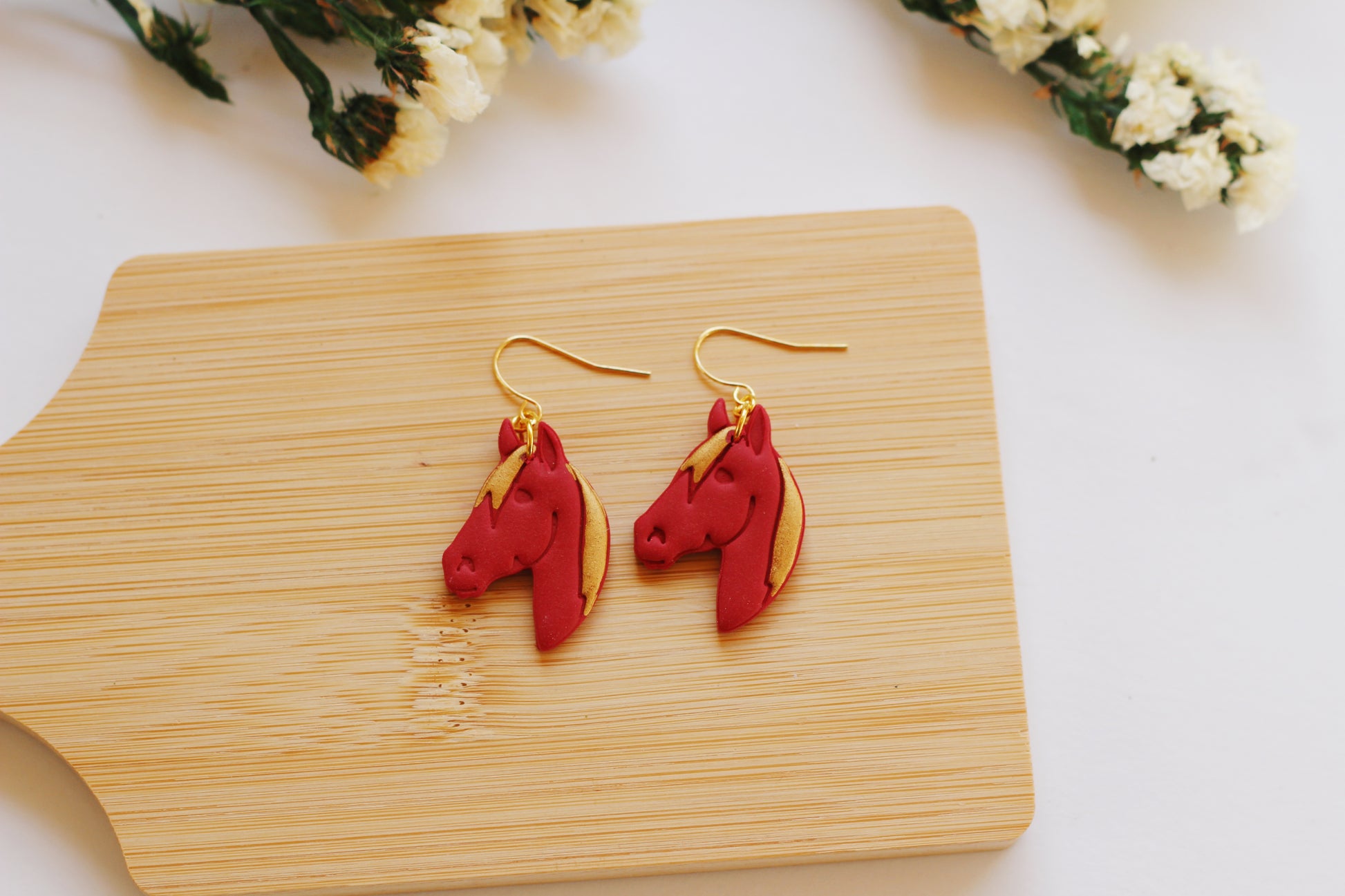 Red fire horse-shaped earrings on a wooden board with flowers in the background