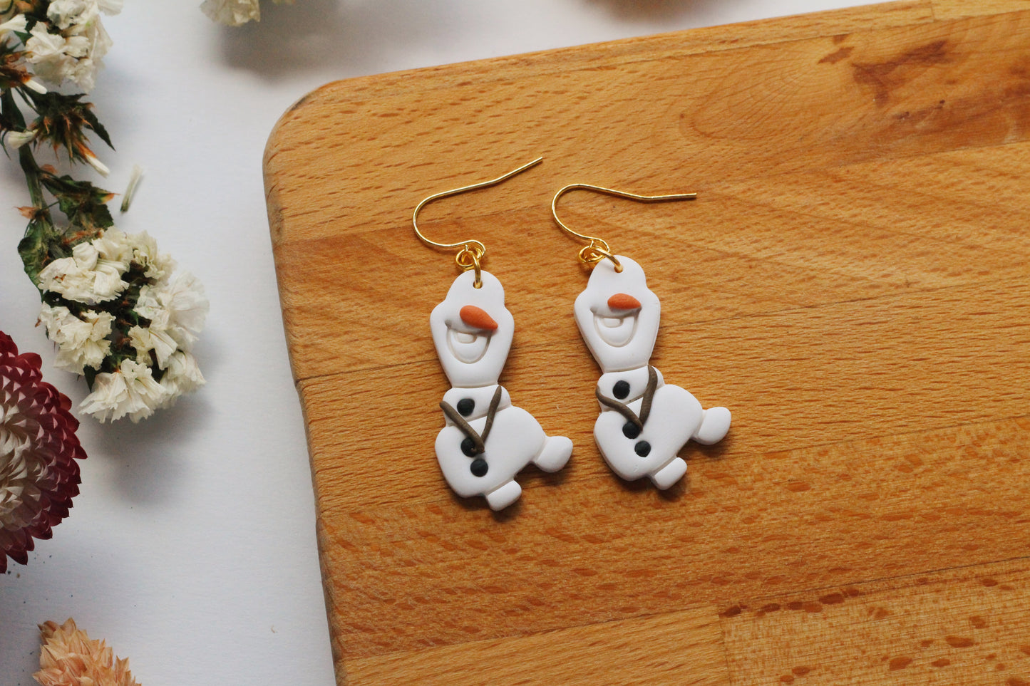 Snowman-shaped earrings on a wooden board with flowers in the background