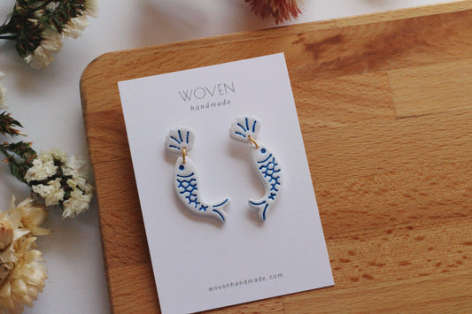 Pair of blue and white earrings on a 'Woven Handmade' card, placed on a wooden surface with flowers in the background.
