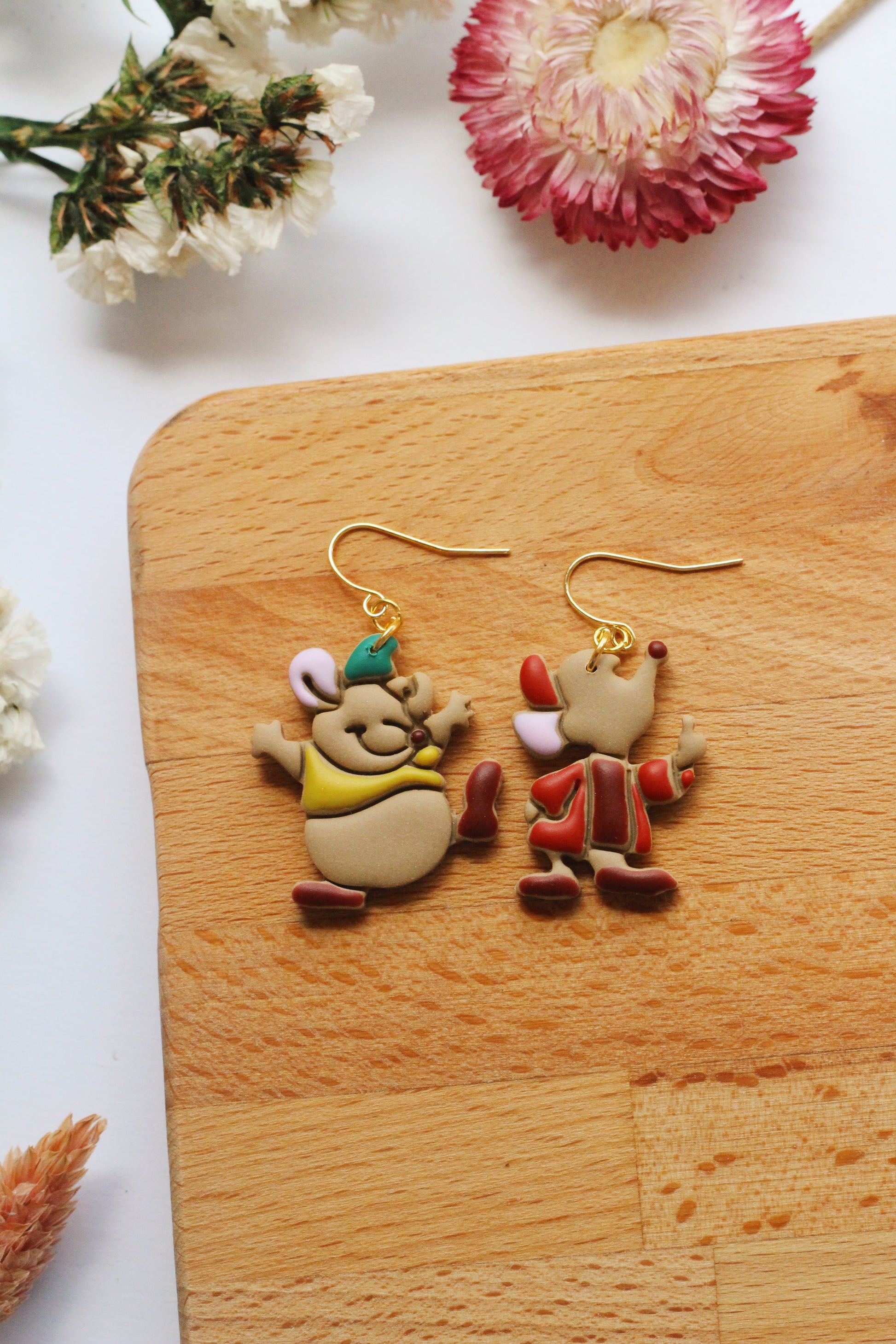 Two cartoon-themed earrings on a wooden board with flowers in the background