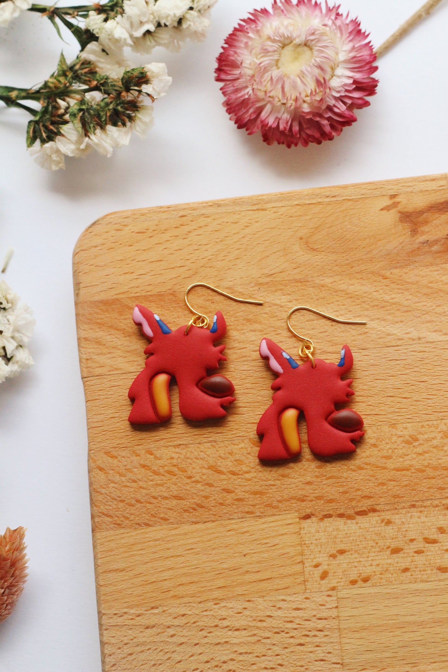 Red dragon-shaped earrings on a wooden board with flowers in the background