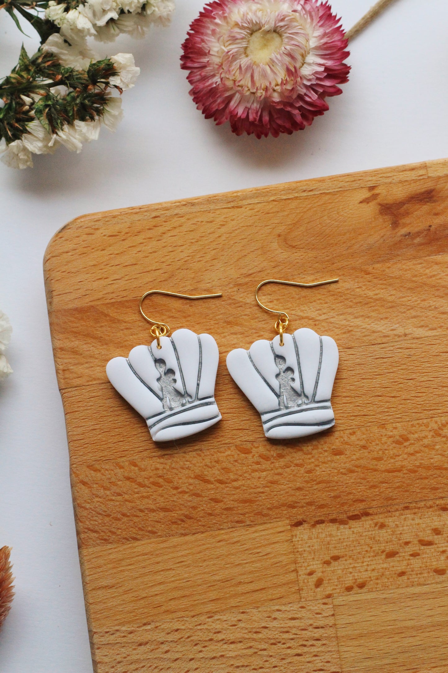 White chef hat earrings with gold hooks on a wooden surface with flowers.