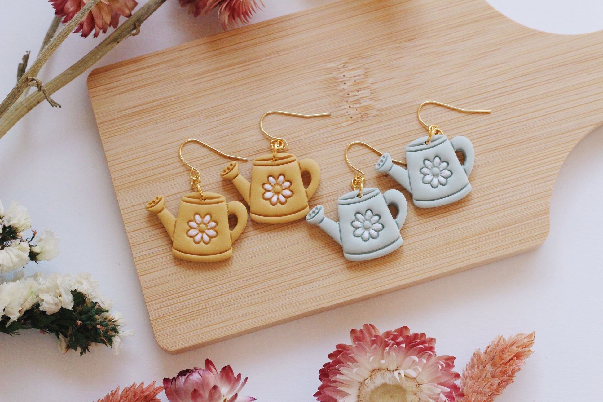 Watering can-shaped earrings on a wooden board with dried flowers around.