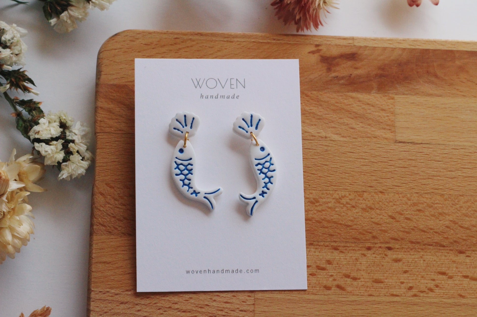 Pair of fish-shaped earrings on a 'Woven Handmade' card with flowers in the background.