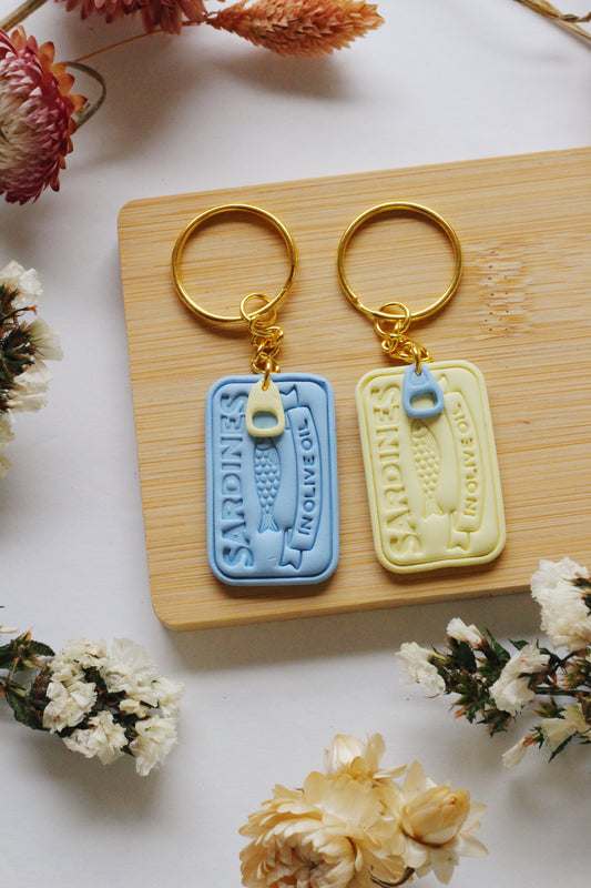 Two sardine can keychains on a wooden board surrounded by flowers