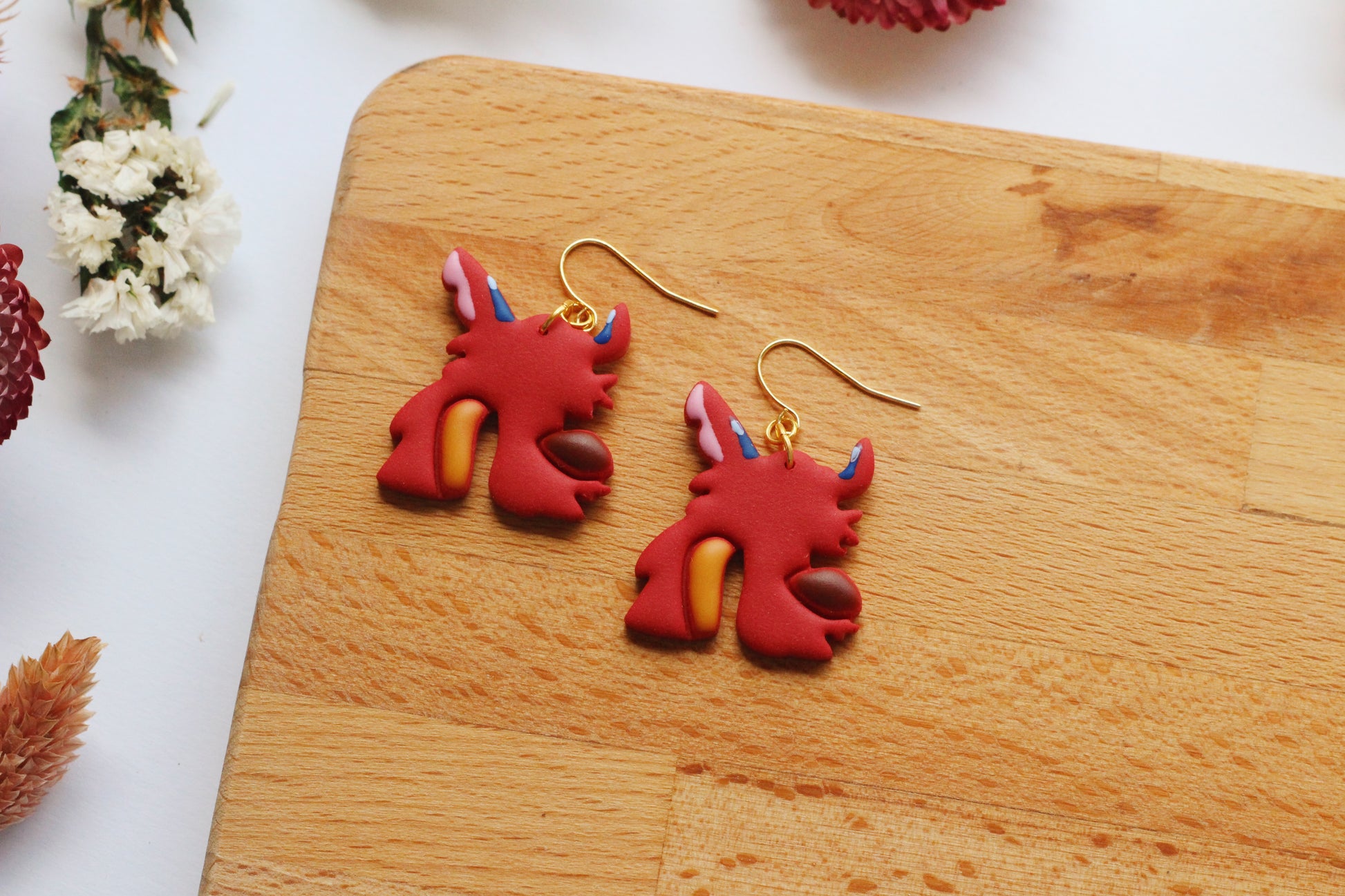 Red dragon-shaped earrings on a wooden board with flowers in the background